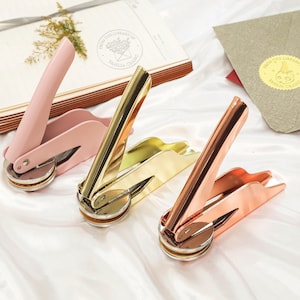 May include: Three gold-toned wax seal stamps with handles. The stamps are in pink, gold, and copper. The stamps are on a white surface.