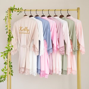 Könnte beinhalten: Ein Set aus acht Roben in verschiedenen Farben, darunter rosa, weiß und grün, mit Spitzenbesatz. Die Roben hängen an einem goldenen Gestell. Die Robe vorne trägt den Namen "Kelly" und das Wort "bridesmaid" ist auf der Rückseite gestickt.