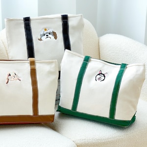 May include: Three canvas tote bags with embroidered dog portraits. The bags have different coloured accents: black, brown, and green. The bags are on a white, cushioned chair.