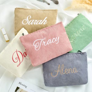 Può includere: Cinque trousse per cosmetici in velluto a coste beige, rosa, verde, grigio e bianco sporco, ciascuna ricamata con un nome in un colore a contrasto. Pennelli per trucco e un rossetto sono visibili sullo sfondo. Le borse sono disposte su una superficie bianca.