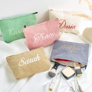 Op de afbeelding: Een verzameling corduroy make-up tassen in verschillende kleuren, waaronder groen, roze, beige, wit en blauw. Elke tas is geborduurd met een naam in een contrasterende kleur. Make-up borstels en compacts zijn zichtbaar in de buurt van een open tas.