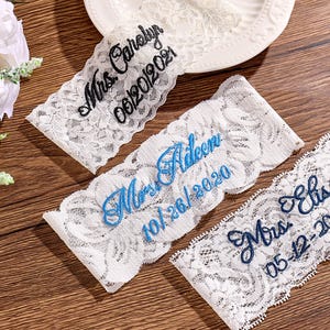 May include: Three white lace garters with embroidered text. One garter has black text reading "Mrs. Carolyn 06/20/2021". Another has blue text reading "Mrs. Aileen 10/26/2020". The third has blue text reading "Mrs. Elisa 05-12-2024".