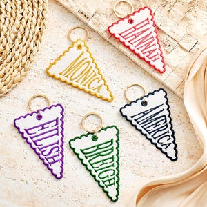 May include: Five triangular white fabric keychains with scalloped edges and gold-tone metal rings. Each keychain features embroidered names in different colours: red for Hannah, yellow for Monica, purple for Eilish, green for Breigh, and navy blue for America.