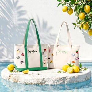 Peut inclure: Deux sacs fourre-tout en toile avec des bordures vertes et crème. Le sac de gauche porte le nom Marlene et des écussons brodés dont une bague, une étoile de mer et un nœud. Le sac de droite porte le nom Carolyn et des motifs comme un flamant rose.