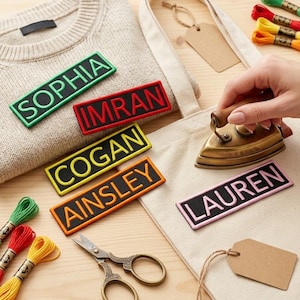 以下が含まれることがあります： SOPHIA、IMRAN、COGAN、AINSLEY、LAURENという名前が刺繍された長方形のワッペン。黒い背景に色付きの縁取りと文字が特徴です。小さな真鍮製のアイロン、金属製のハサミ、色とりどりの刺繍糸が一緒に置かれています。