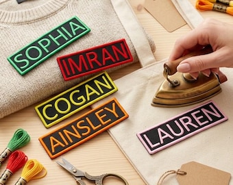 Op de afbeelding: Rechthoekige geborduurde naamlabels met de namen SOPHIA, IMRAN, COGAN, AINSLEY en LAUREN. De labels hebben gekleurde randen en letters op een zwarte achtergrond. Een klein koperen strijkijzer, een metalen schaar en borduurgaren zijn aanwezig.