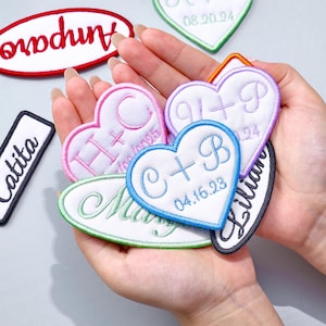Pode incluir: Uma coleção de patches bordados em várias formas, incluindo corações e ovais. Os patches apresentam texto personalizado em diferentes cores, como rosa, azul e verde, com datas e iniciais. Os patches são brancos com bordas coloridas.