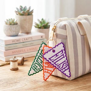 May include: Three triangular felt pennant keychains with embroidered names Eilish, Savannah, and Raleigh in purple, orange, and green thread, attached to a gold metal keyring on a beige and white striped canvas zipper pouch.