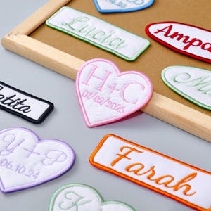 Könnte beinhalten: Verschiedene gestickte Aufnäher in verschiedenen Formen, darunter Rechtecke und Herzen. Die Aufnäher zeigen Namen und Initialen in verschiedenen Farben wie Grün, Orange und Rosa mit Daten. Die Aufnäher werden auf einem Holzrahmen präsentiert.