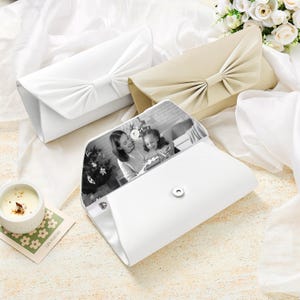 May include: Three rectangular clutch bags with a pleated bow design on the front flap. One bag is white, one is beige, and one is white with an open flap revealing a black and white photograph of a person holding a child inside the interior lining.
