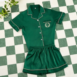May include: A green satin pajama set with short sleeves and shorts. The top has a white collar and a pocket with an embroidered letter "G" surrounded by a wreath.