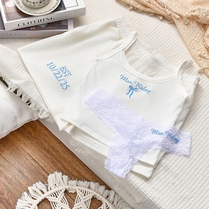 May include: White bridal lingerie set featuring a tank top and a matching bag, both embroidered with blue text. The tank top has "Mrs. Riley" embroidered on it, and the bag has "EST 10/22/25". A white lace thong is also included.