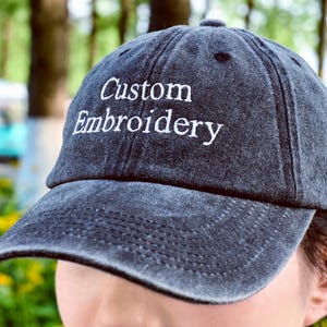 Può includere: Un cappellino da baseball grigio scuro con le parole "Custom Embroidery" ricamate in bianco. Il cappellino ha una visiera curva e un aspetto classico e usurato. Lo sfondo è una scena all'aperto sfocata.