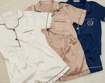 May include: Three sets of satin pyjamas in white, blush pink, and navy blue. Each set includes a short-sleeved button-down shirt and matching shorts, trimmed with black piping. The shirts feature a pocket with a gold embroidered initial.