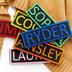 May include: Embroidered name patches in various colours, including red, yellow, green, blue, orange, and pink. Each patch features a black background with a name stitched in a contrasting colour. The patches are rectangular and have a border.