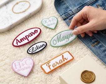 May include: A collection of embroidered fabric patches featuring names and dates, including heart-shaped, oval, and rectangular designs. The patches display text such as Amparo, Lilian, Maryse, Farah, K+E 08.20.24, and H+C 07/02/2025 in various thread colors.