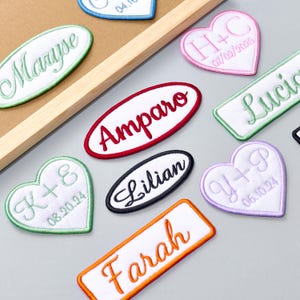 Puede incluir: Parches bordados de diversas formas, incluyendo óvalos, corazones y rectángulos. Cada parche presenta un nombre o iniciales en un color contrastante, como "Maryse", "Amparo" y "Lilian". Los parches tienen un fondo blanco con bordes coloridos.