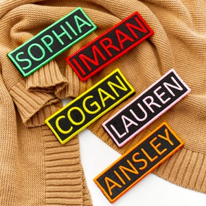 May include: A collection of embroidered name patches in different colours, including Sophia, Imran, Cogan, Lauren, and Ainsley. The black patches have coloured borders and lettering, displayed on a tan knitted jumper.