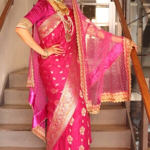 May include: A vibrant pink sari with gold embroidery and floral patterns. The ensemble includes a matching veil with gold trim, a velvet blouse, and ornate gold jewelry. The outfit is worn with black heels, creating a striking and elegant look.