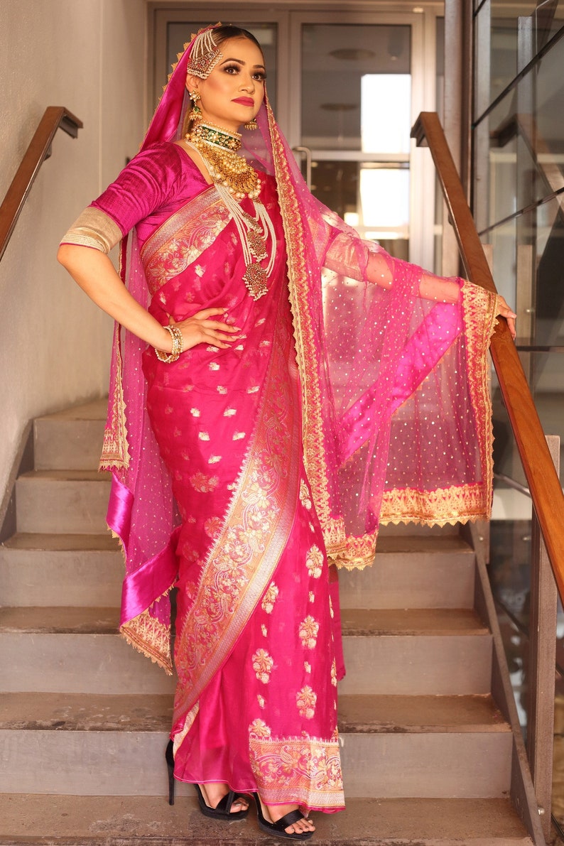 May include: A vibrant pink sari with gold embroidery and floral patterns. The ensemble includes a matching veil with gold trim, a velvet blouse, and ornate gold jewelry. The outfit is worn with black heels, creating a striking and elegant look.