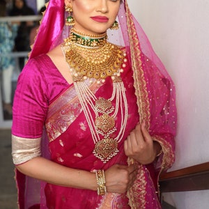 May include: A woman in a magenta sari and matching veil, adorned with elaborate gold jewelry. The jewelry includes a choker, layered necklaces, earrings, a headpiece, and bracelets. The sari features gold embroidery.