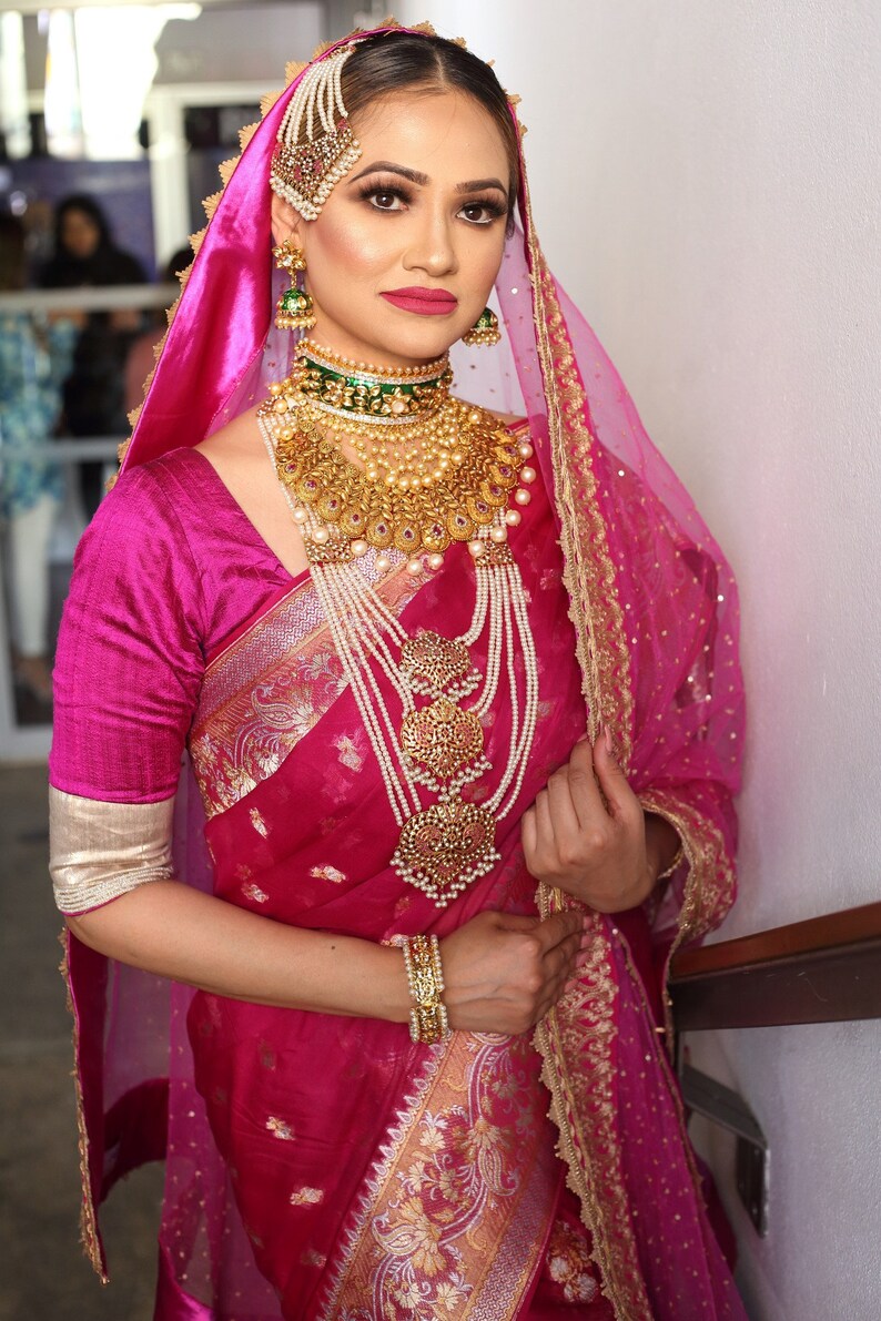 May include: A woman in a magenta sari and matching veil, adorned with elaborate gold jewelry. The jewelry includes a choker, layered necklaces, earrings, a headpiece, and bracelets. The sari features gold embroidery.