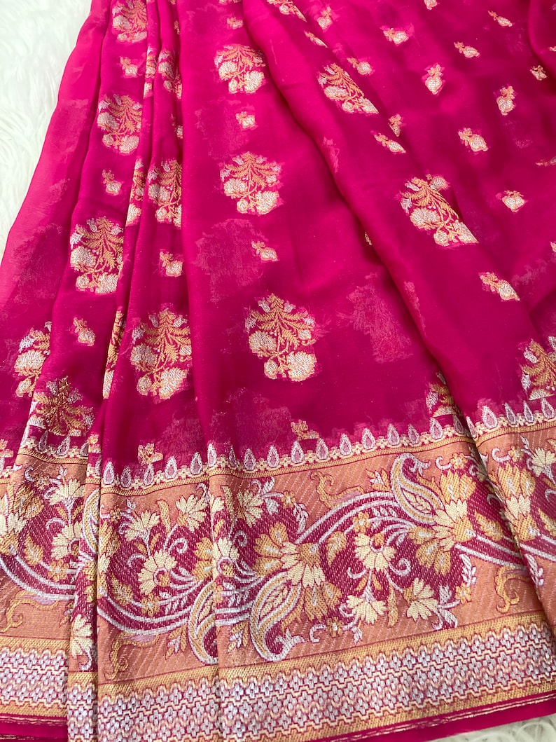 May include: A vibrant fuchsia saree with gold floral and paisley patterns. The sheer fabric drapes elegantly, showcasing intricate details along the border. This traditional garment is perfect for special occasions.