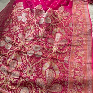 May include: A vibrant fuchsia-colored textile with an intricate gold and silver floral pattern. The fabric features a detailed border with similar floral designs. The overall aesthetic suggests a luxurious garment or decorative piece.
