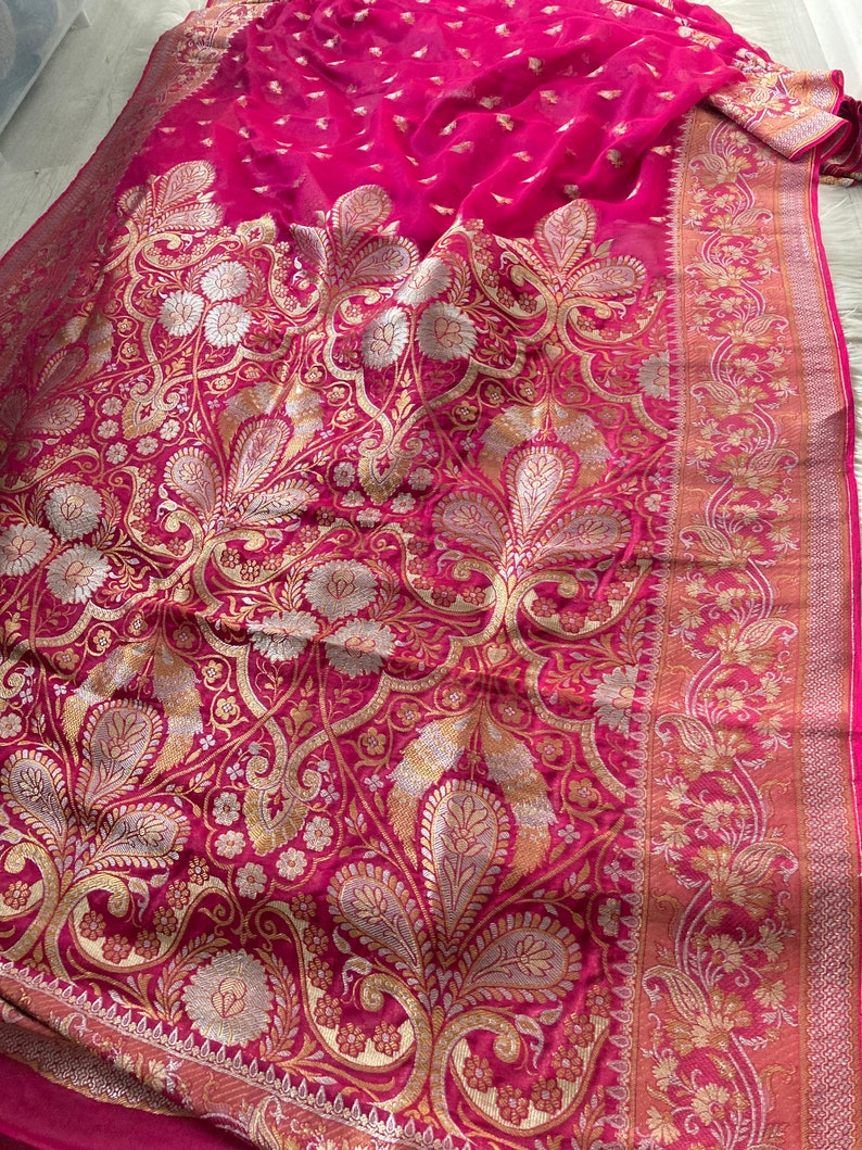 May include: A vibrant fuchsia-colored textile with an intricate gold and silver floral pattern. The fabric features a detailed border with similar floral designs. The overall aesthetic suggests a luxurious garment or decorative piece.