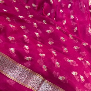 May include: A vibrant magenta fabric with a delicate, sheer texture. The material is adorned with scattered gold-colored floral patterns. A decorative border with a silver and gold geometric design runs along one edge, adding a touch of elegance.