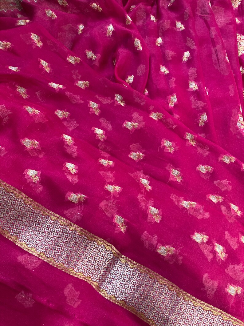 May include: A vibrant magenta fabric with a delicate, sheer texture. The material is adorned with scattered gold-colored floral patterns. A decorative border with a silver and gold geometric design runs along one edge, adding a touch of elegance.