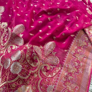 May include: A vibrant fuchsia textile with intricate gold and silver paisley and floral patterns. The fabric has a delicate, sheer quality, with a detailed border. The design features small, scattered gold accents throughout the material.