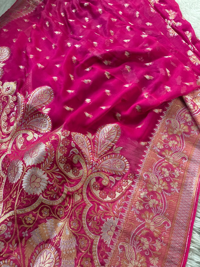 May include: A vibrant fuchsia textile with intricate gold and silver paisley and floral patterns. The fabric has a delicate, sheer quality, with a detailed border. The design features small, scattered gold accents throughout the material.