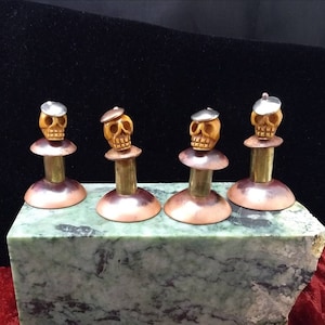 May include: Four small, yellow skull figurines with silver hats are perched on top of copper and brass bases. The bases are arranged in a row on a green marble surface.