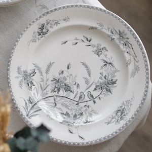 Puede incluir: Plato de cerámica blanca con un diseño floral y de pájaros detallado en tonos grises. El plato tiene un borde decorativo y está colocado sobre un mantel de tonos neutros. Un segundo plato es parcialmente visible en el fondo.
