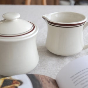 Puede incluir: Azucarero de cerámica color crema con tapa y jarra de crema con una raya marrón oscuro. El azucarero es redondo con tapa y un pequeño pomo. La jarra de crema tiene asa y pico. Un libro está en primer plano.