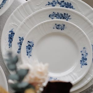 Puede incluir: Una pila de platos de porcelana blanca con un diseño floral azul. Los platos, de diferentes tamaños, presentan un delicado patrón en relieve en los bordes. Los detalles florales azules añaden un toque de elegancia a la vajilla.