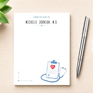 May include: A white notepad featuring the text FROM THE DESK OF MICHELLE JOHNSON, M.D. in blue ink. The design includes a small blue heart, a watercolor illustration of a medical clipboard with a red heart, and a blue stethoscope at the bottom.