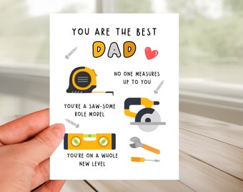 Puede incluir: Una tarjeta de felicitación blanca con el texto "YOU ARE THE BEST DAD" e ilustraciones de herramientas. La tarjeta incluye una cinta métrica, una sierra, un nivel, una llave inglesa y un destornillador, con las frases "NO ONE MEASURES UP TO YOU" y "YOU'RE ON A WHOLE NEW LEVEL."