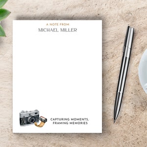 May include: A white notepad with a gold border and the text "A Note From Michael Miller" at the top. The bottom of the notepad features a watercolor illustration of a vintage camera with a roll of film. The text "Capturing Moments, Framing Memories" is below the illustration.