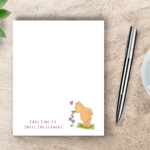 May include: A white notepad with a cartoon bear smelling flowers and a butterfly. The notepad has the text "Take time to smell the flowers" printed on it.
