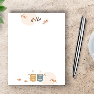 May include: A white notepad with the word "Hello" at the top and a design of two coffee mugs with the text "Stay Cozy" and "Stay Happy" at the bottom. The notepad is on a wooden surface with a pen and a cup of coffee.