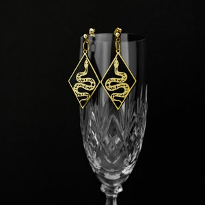 May include: A pair of gold-tone earrings with black enamel snake designs. The earrings are hanging from a crystal glass.