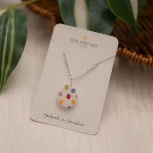 以下が含まれることがあります： カラフルな水玉模様で飾られた卵型のペンダントが付いたシルバーネックレス。ペンダントは「SUN AND SKY」と「Handmade in Lancashire」と書かれた白いカードに表示されています。