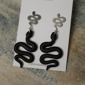 May include: A pair of black snake earrings with silver snake accents. The earrings are dangling and have a simple design.