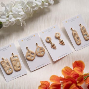 May include: Four pairs of beige polymer clay earrings displayed on white cards labeled SUN AND SKY DESIGNS. The earrings feature gold-tone posts and various shapes including rectangles, teardrops, and shell-like designs with embedded translucent accents.
