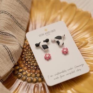 May include: A pair of stud earrings shaped like black and white cow heads and a pair of small pink flower stud earrings on a white display card labeled SUN AND SKY. The card includes text stating the items are sustainably made from recycled clay.