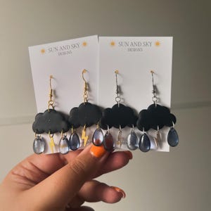 May include: A pair of black cloud earrings with silver and gold lightning bolt charms and blue raindrop charms. The earrings are on a white card with the text "SUN AND SKY DESIGNS".