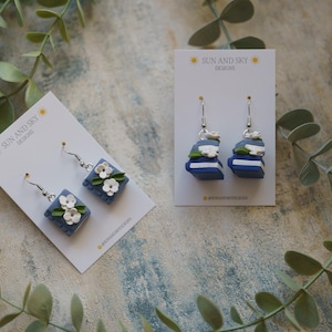 May include: Two pairs of earrings featuring blue and white clay book and flower designs. The earrings are on white cards with the text "Sun and Sky Designs".
