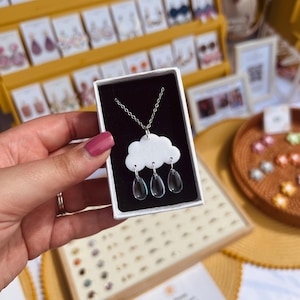 May include: A silver chain necklace with a white ceramic cloud pendant and three clear glass raindrops hanging from it. The necklace is displayed in a small white box.